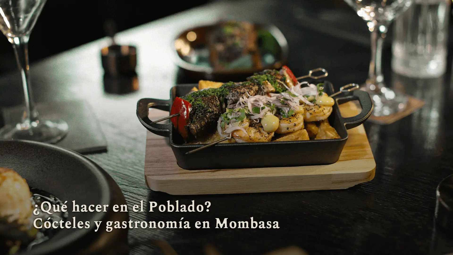 Qué hacer en El Poblado Cast-iron skillet with grilled skewers, potatoes and onions on a wooden board; restaurant setting with the Spanish headline: ¿Qué hacer en el Poblado? Cócteles y gastronomía en Mombasa.