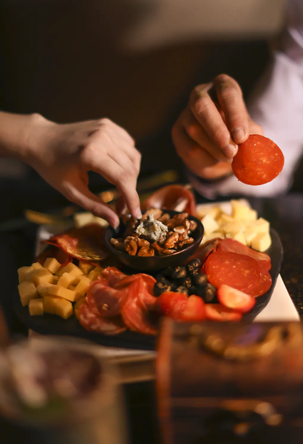 Charcuterie board with cubed cheese, sliced salami, walnuts, olives, and berries; a person adds a piece of meat on the board.