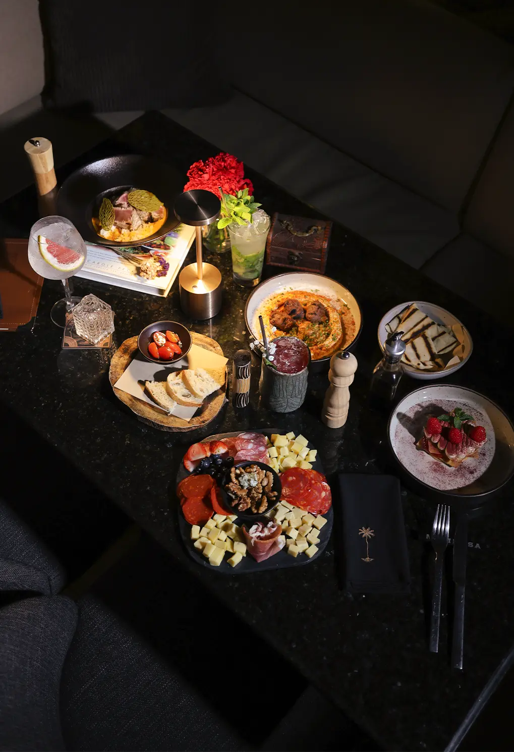 Charcuterie board with cheeses, cured meats, bread and nuts on a dark table, with cocktails nearby.