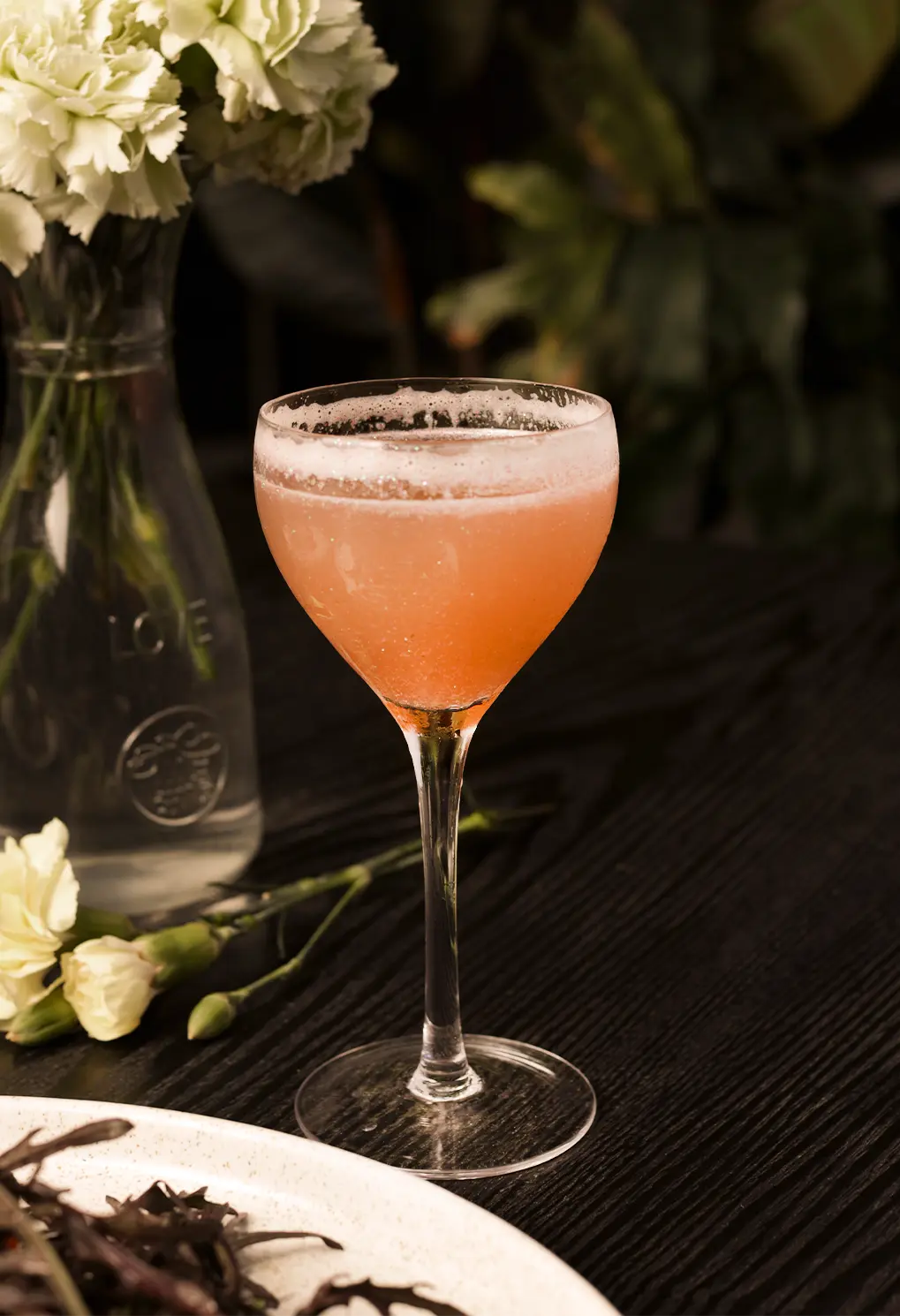 Pink cocktail in a coupe glass on a dark wooden table, with a vase of white flowers beside it.