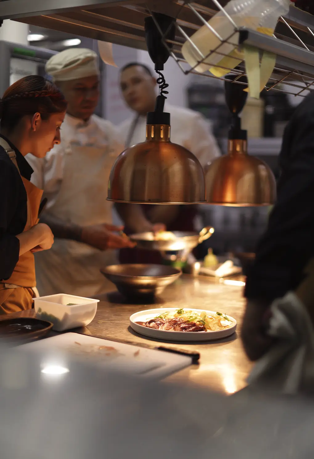 Chef plating a dish under warm copper lamps in a busy kitchen setting.
