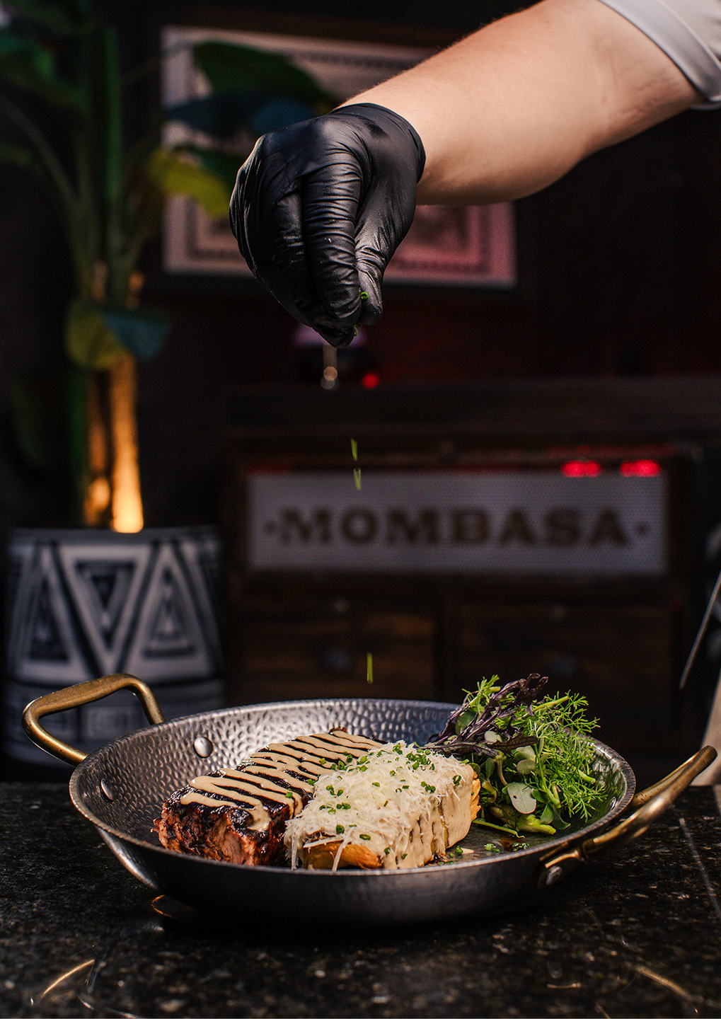 Chef with gloved hand sprinkling herbs over a plated steak with salad in a hammered pan, ready to serve in a restaurant kitchen.