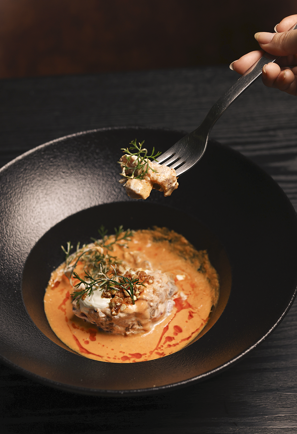 Gourmet dish in a black bowl with creamy orange sauce and thyme garnish; a fork lifts a bite-sized piece above the plate.