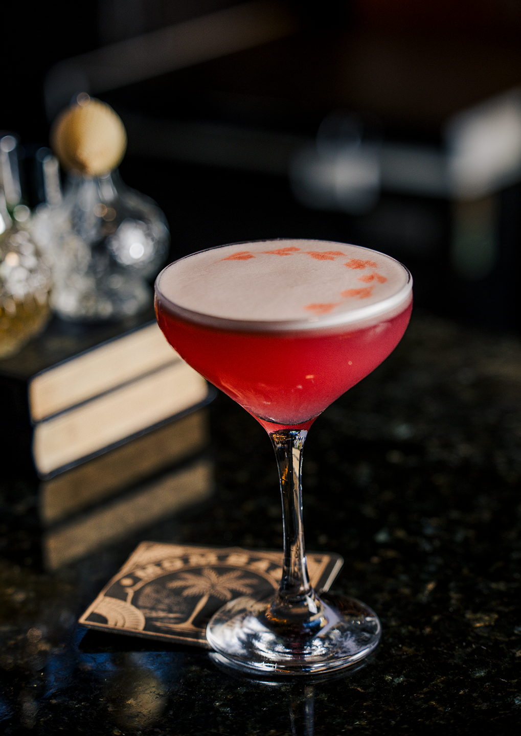 Pink cocktail in a coupe glass with a foamy top on a dark bar counter, blurred bottles in the background.