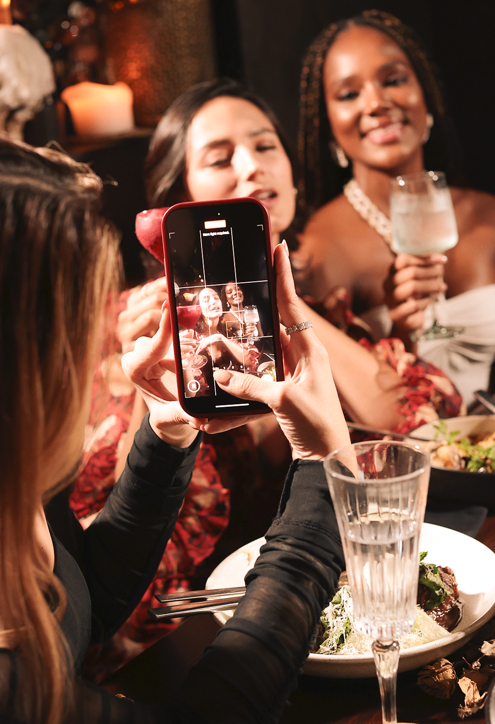 Group of friends at a dinner table posing for a photo with a smartphone; one person holds the phone while two women smile in the background, drinks and plates visible.