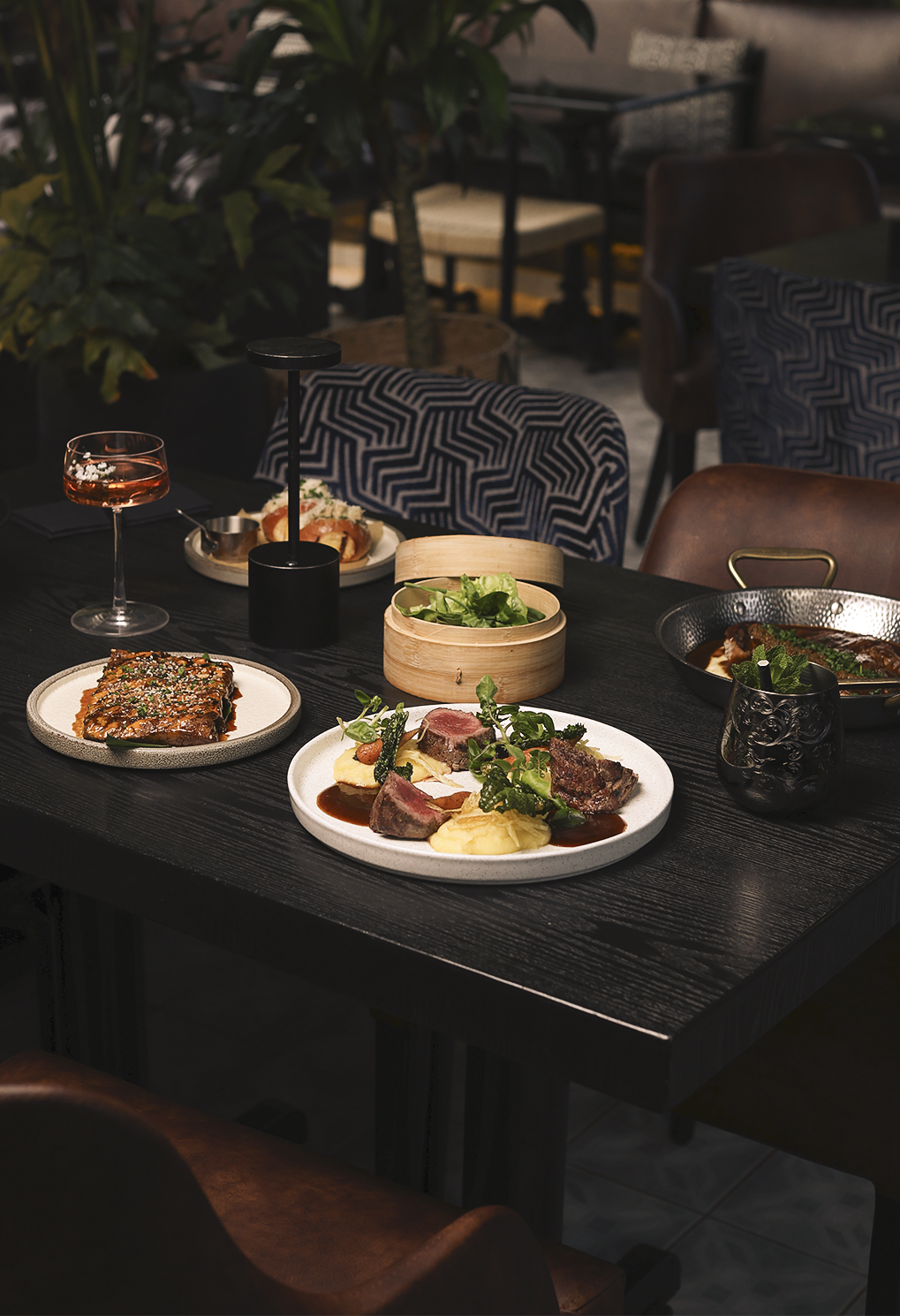 A dark wooden dining table with several plated dishes: steak, greens, and side items in a stylish restaurant setting.