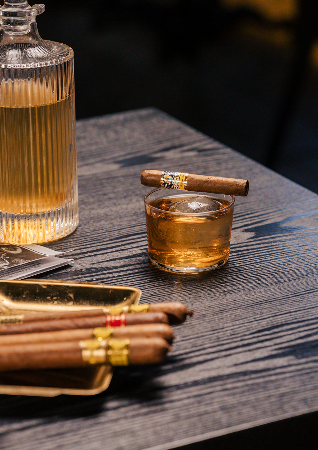 Whiskey on the rocks in a short glass with a cigar balanced on the rim, on a dark wooden table beside a glass decanter and cigars in a tray.
