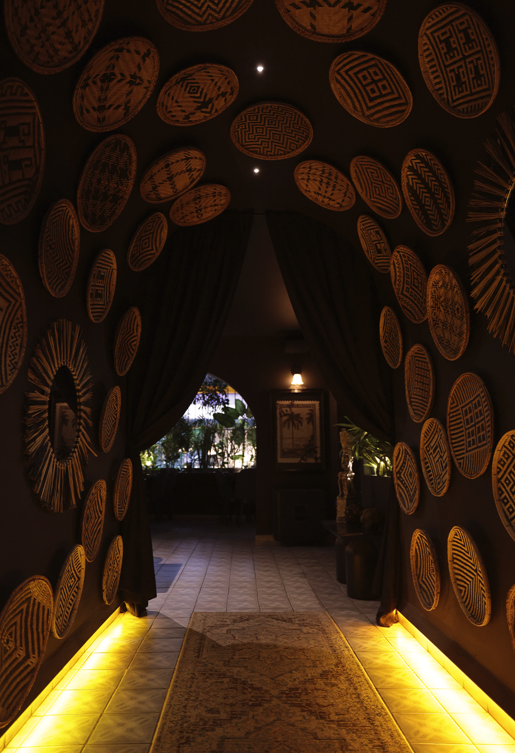 Dark hallway lined with woven baskets on the walls and yellow floor lighting guiding toward a room with plants beyond an arched doorway.