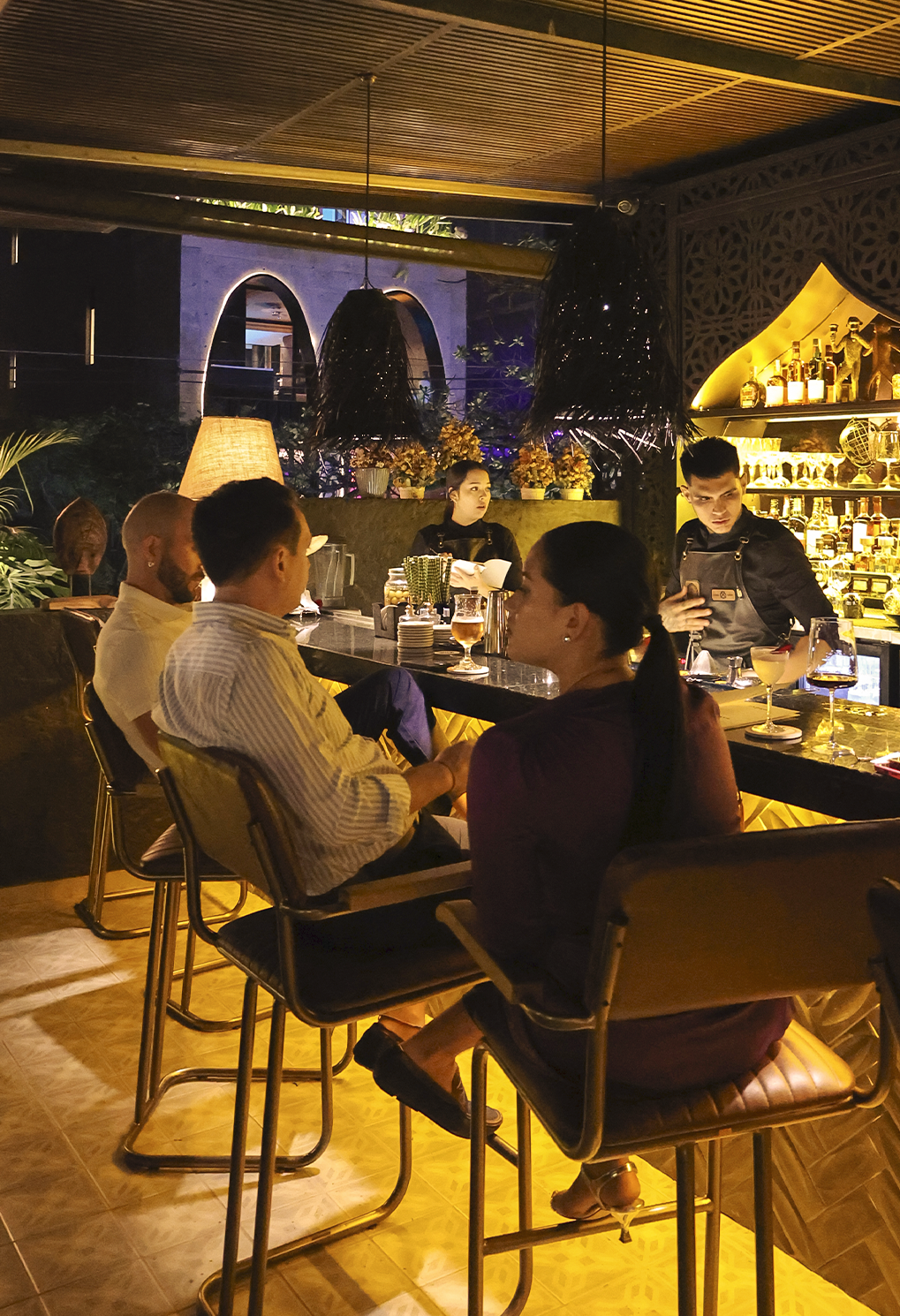 People seated at a bar counter in a warmly lit restaurant, bartenders mix drinks in the background.