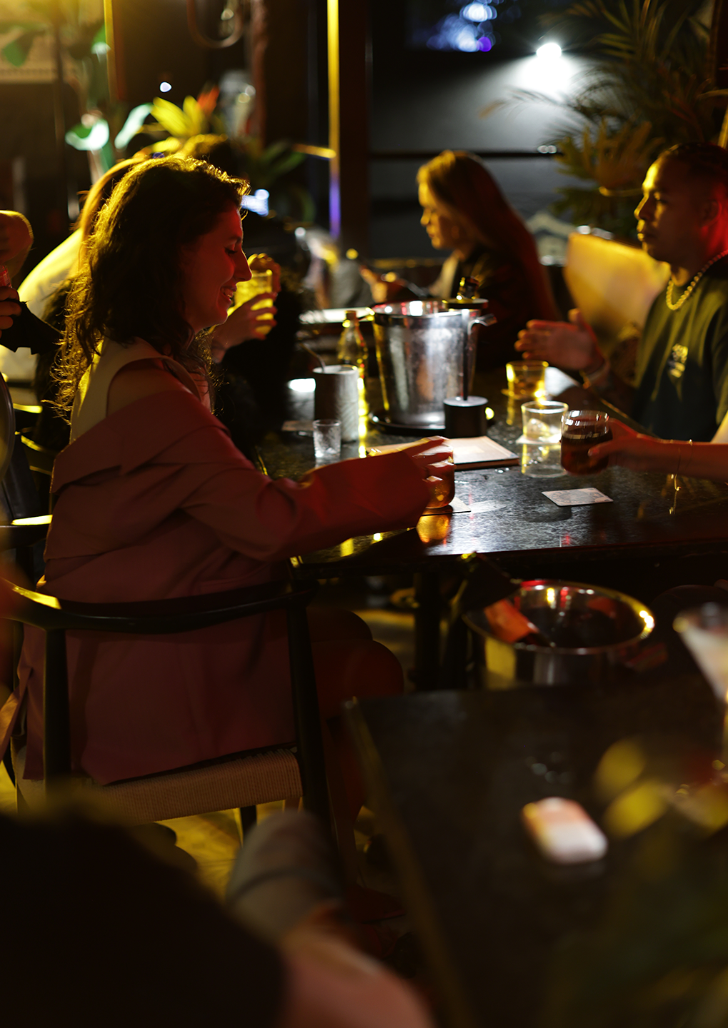 Woman in a pink coat sits at a bar smiling while talking to friends in a dim, colorful-lit club with drinks on the table.