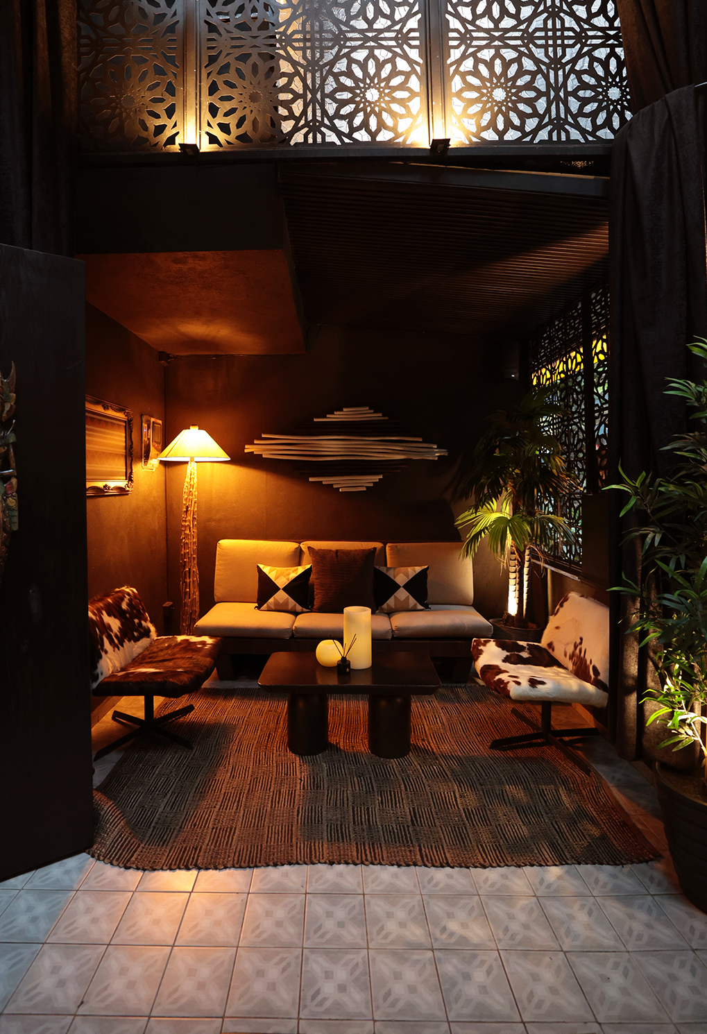 Cozy living room lounge with a beige sofa, cowhide chairs, warm lighting, and a low coffee table on a rug, in a dimly lit interior.