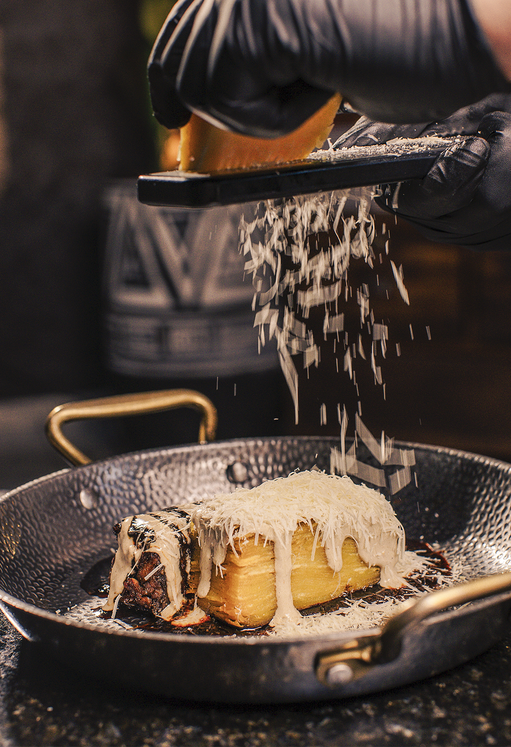 Chef grating cheese over a plated dish in a dark skillet, adding a snow of shredded cheese as it’s prepared tableside