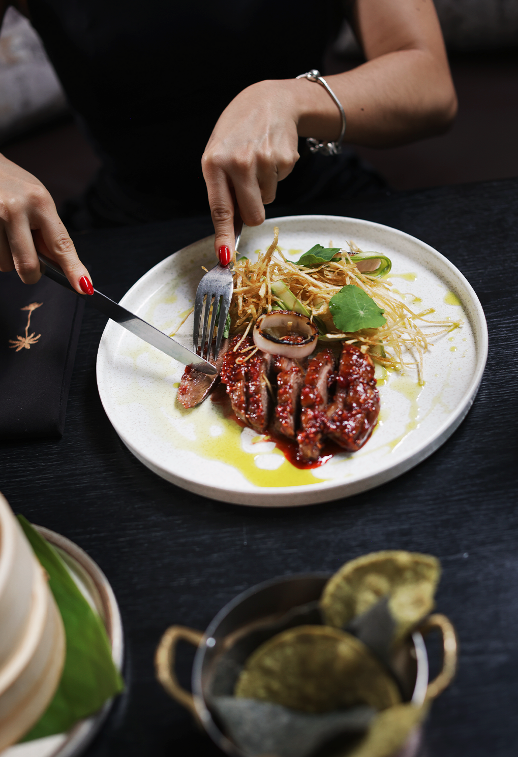 Hands cut into a plated beef dish with caramelized slices, crispy garnish, and greens on a white plate at a dark table.