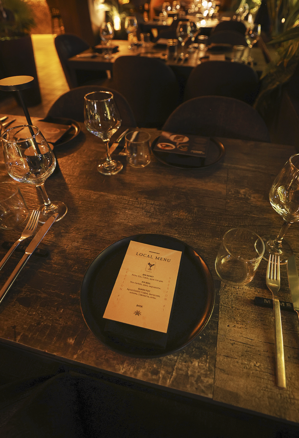Table set in a dim restaurant with a black plate and local menu card at center, surrounded by wine glasses and silverware on a rustic wooden table