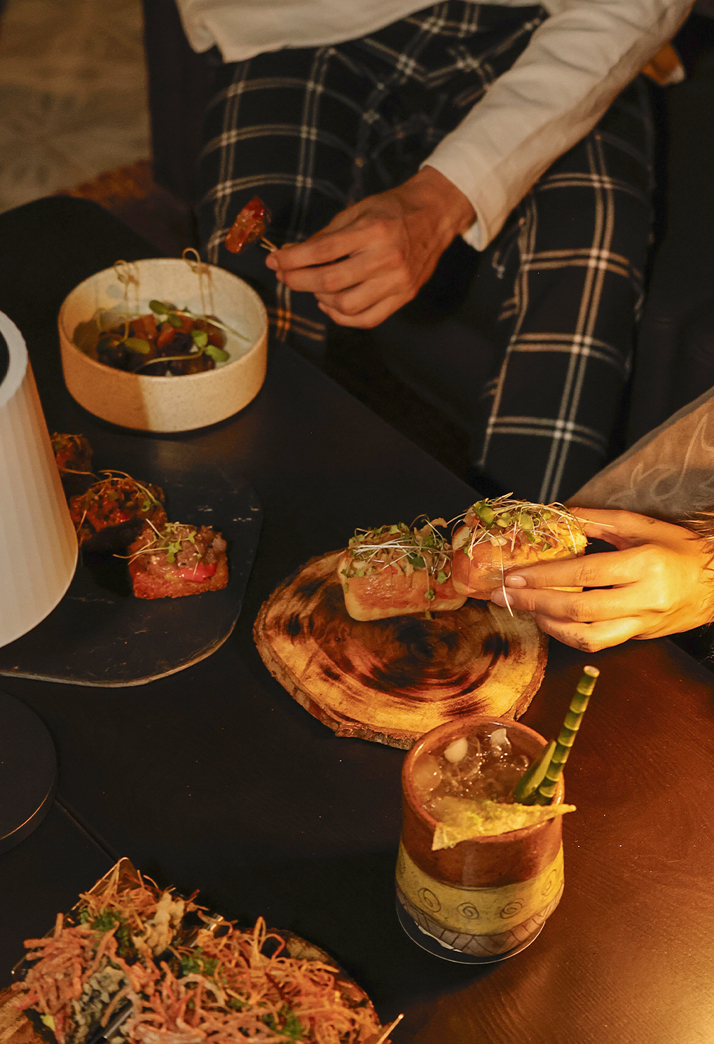 A person places a herb-topped canapé on a rustic wooden board while other bites and a drink sit on a dark table.